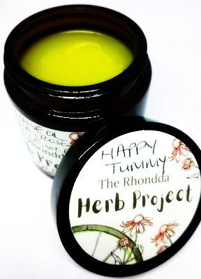 Open jar of yellow balm labeled 'Happy Tummy Herb Project' with botanical illustrations.